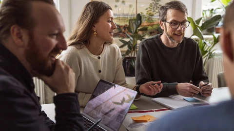 Et gruppe mennesker deltager i en brainstormingsession ved et bord, med en bærbar computer og farvede post-it sedler, omgivet af planter i et lyst rum.